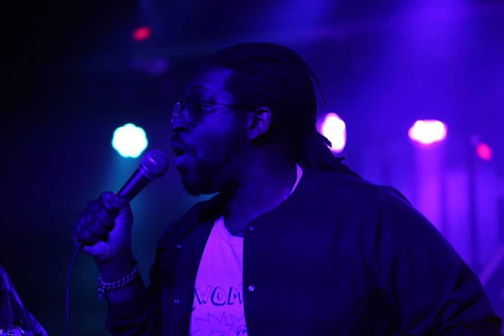 Soul Glo vocalist Pierce Jordan sings into the microphone he is handholding. He is position in profile in the center of the image, looking directly out to the left. He is wearing a button-down shirt that is opened enough to reveal a t-shirt with some partially obscured cartoon drawing and text. He is wearing glasses, a heavy chain bracelet is visible on his right wrist, which is the hand that's supporting the microphone. He has a beard and his locs are pulled back into a ponytail. The entire image is cast in a deep blue hue from the stage lights.
