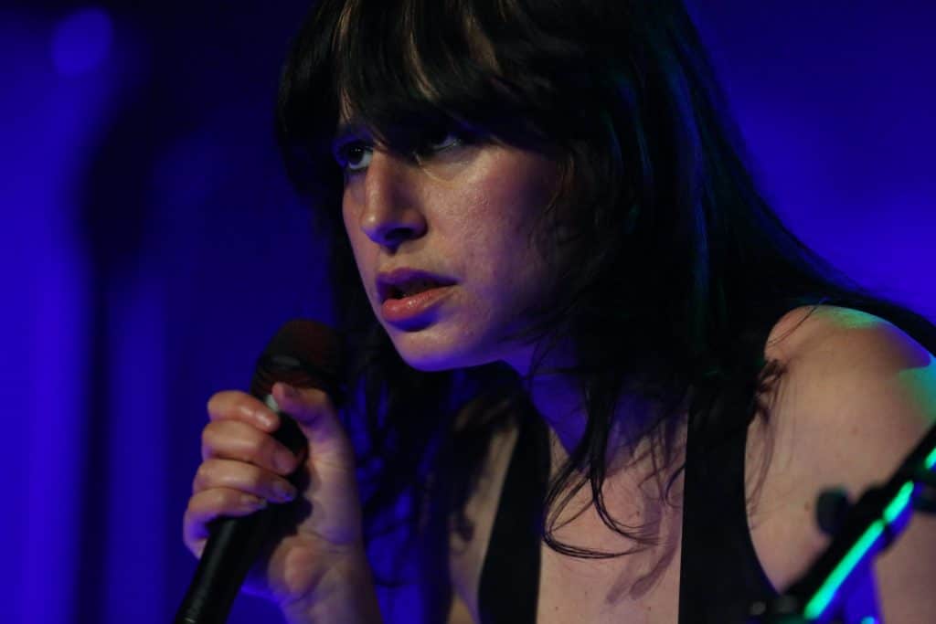Mannequin Pussy guitarist/vocalist Missy Dabice looks out at the audience, with a microphone clutched in her right hand. She is in a tight closeup. Black bangs fall over her eyes and the shoulder straps of her black leather chest bodice are visible. The background is a vivd blue.