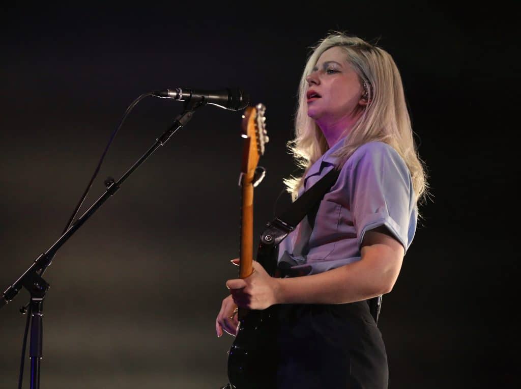 Alvvays guitarist/vocalist Molly Rankin approaches a microphone while looking out over the audience. She's wearing a baby blue button-up t-shirt and black pants. She is in profile, positioned to the slight right of the image, while the microphone is slightly left. The background behind her is dark, but she is back-lit by stage lighting, creating a soft glow effect.