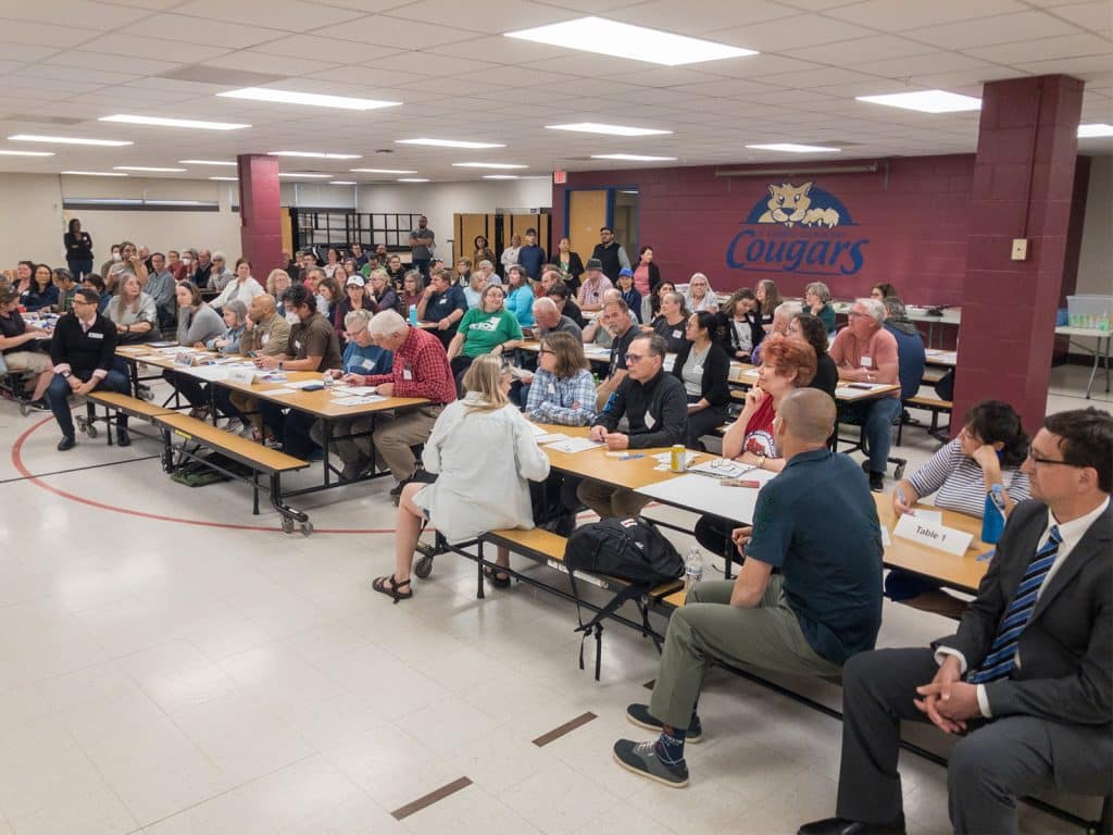 A wide photo of an elementary school cafeteria. Adults fill most of the cafeteria benches with papers on the numbered tables.