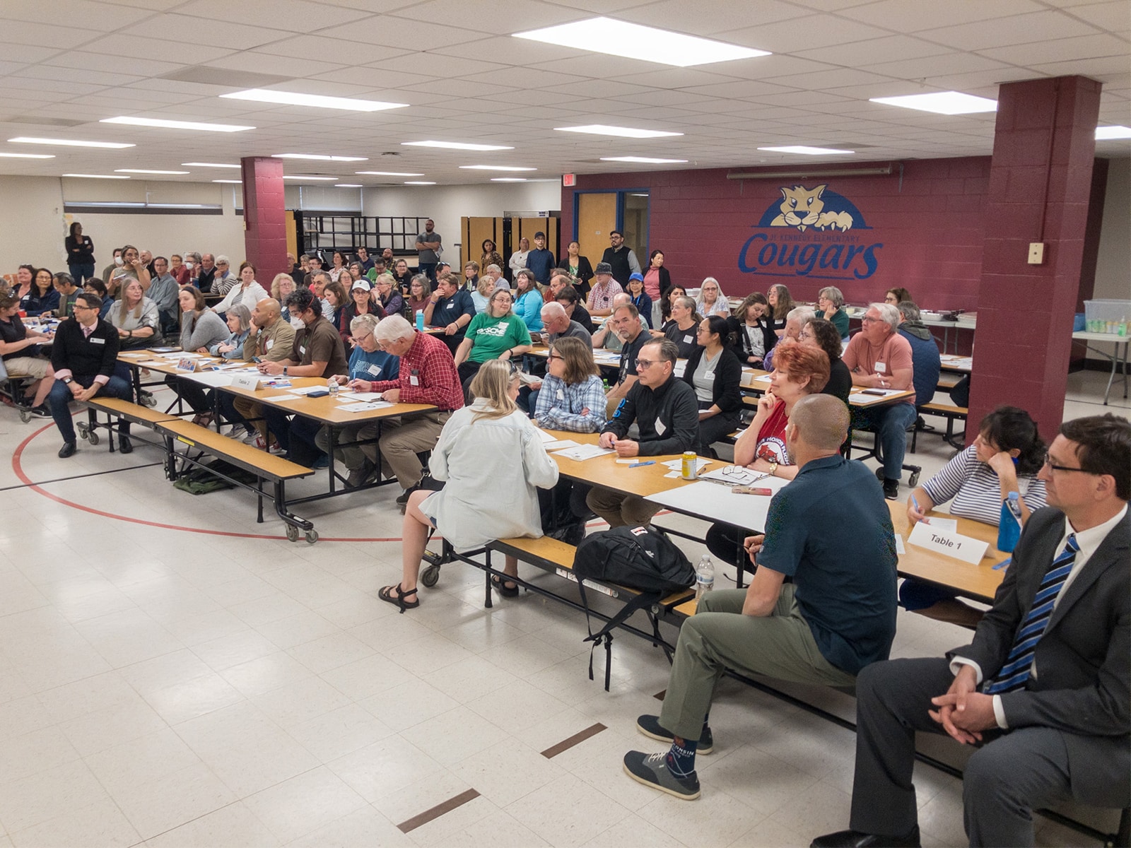 A wide photo of an elementary school cafeteria. Adults fill most of the cafeteria benches with papers on the numbered tables.
