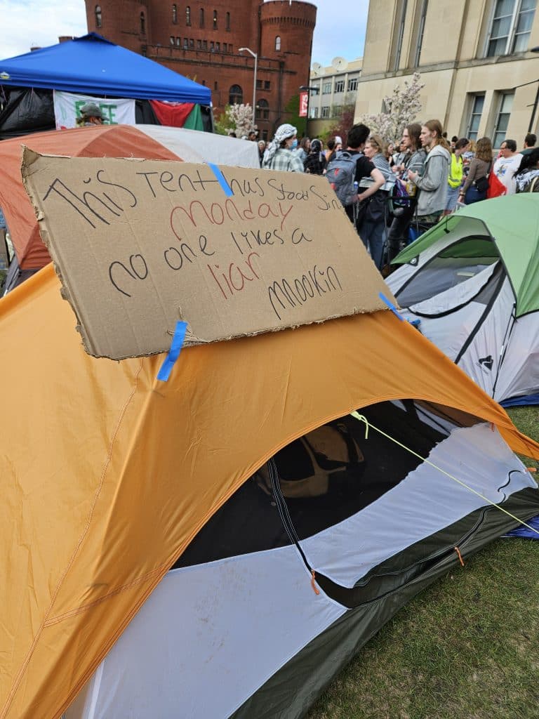 A photo shows a brightly colored tent decorated with a handwritten cardboard sign reading, "This tent has stood since Monday. No one likes a liar, Mnookin." Groups of demonstrators are visible in the background, as are UW-Madison's Red Gym and Memorial Library buildings.