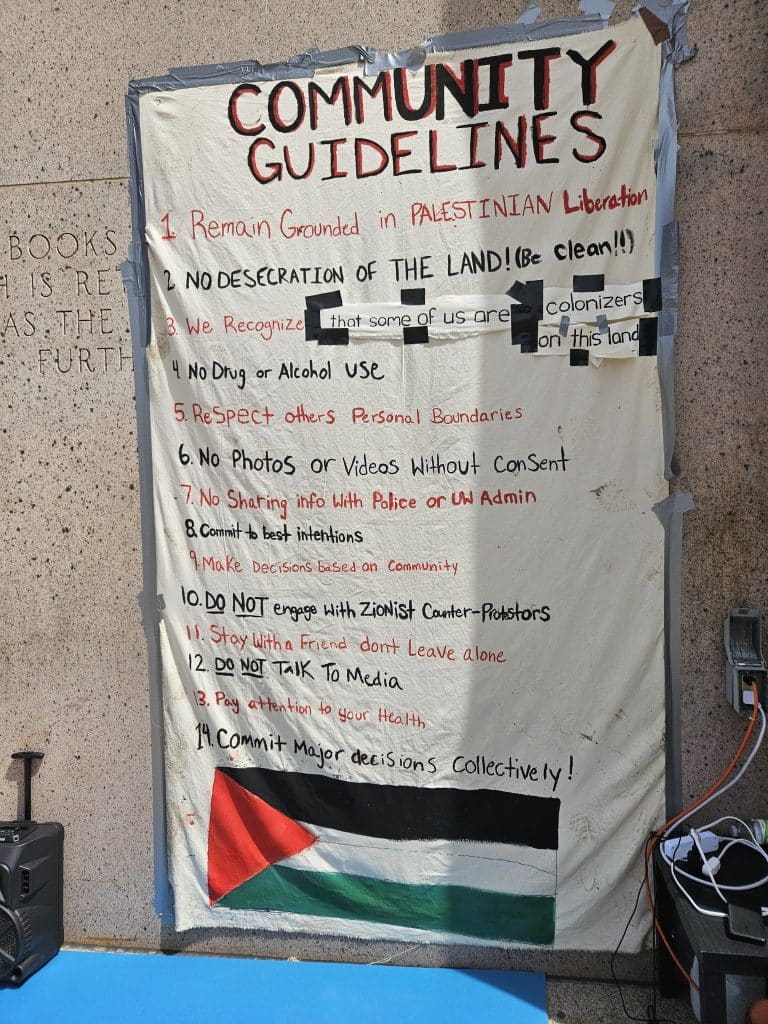 A photo shows a handwritten sign taped to the exterior wall of a building, detailing community guidelines for a protest and decorated with a Palestinian flag.