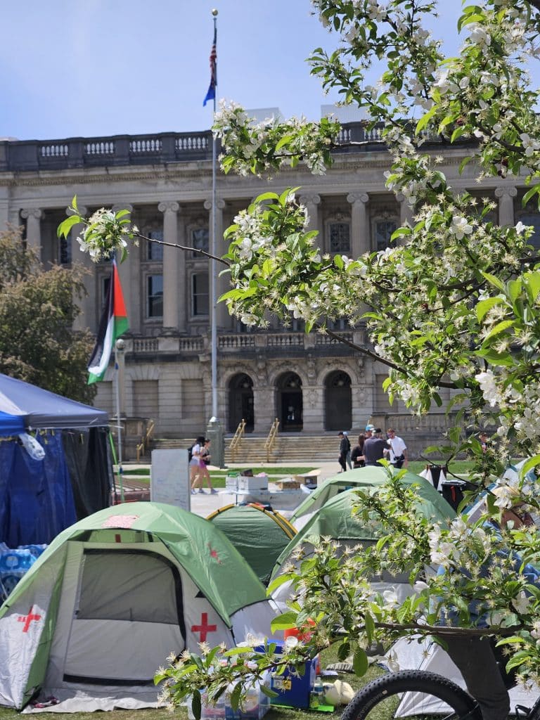 A photo shows a group of green tents on Library Mall, one with visible red cross symbols on it indicating a medic tent. In the foreground are branches from a blooming tree. In the background, a Palestinian flag is visible, as is the Wisconsin State Historical Society building.