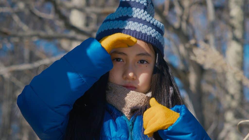 At a close-up, eight-year-old Hana looks out into the distance at something off screen near the woods. She holds her right hand up to her forehead to shield her eyes from the sun. Hana is bundled in winter attire that includes mustard-yellow mittens, a horizontally-striped toque with dark and light blues, tan scarf, and a vivid blue heavy coat.
