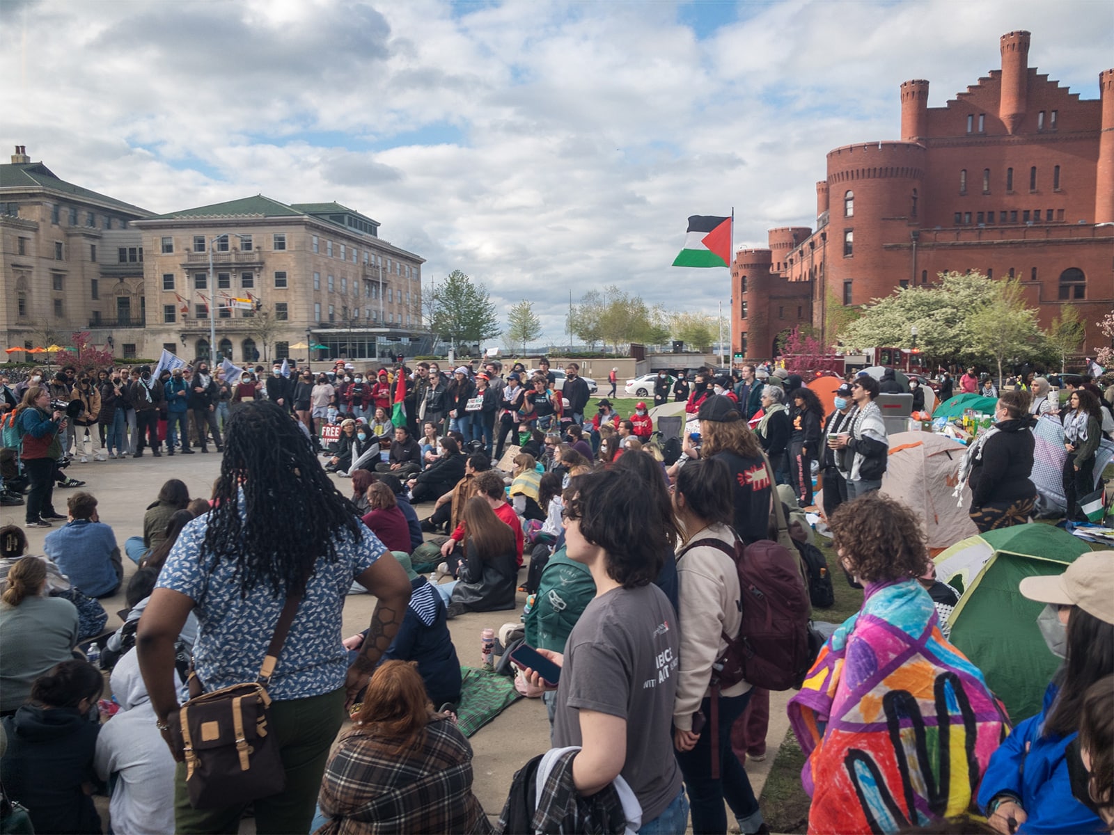 A photo shows a large group of protestors gathered in a circle on UW-Madison’s Library Mall on a sunny afternoon. Behind them, the Memorial Union and Red Gym buildings frame a background view of the sky and Lake Mendota.