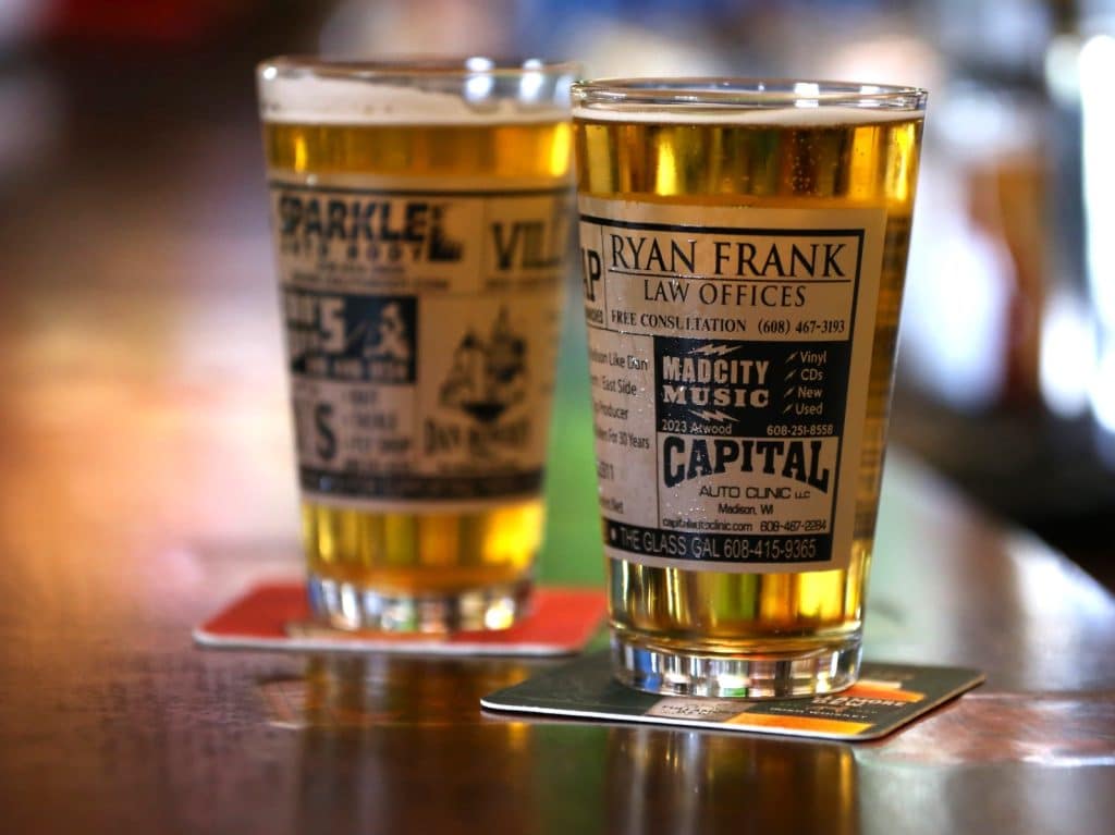 Two pint glasses are shown on the bar of Villa Tap. They are on coasters, one is clearly visible to the near-right of the image, while the glass situated just behind is in soft focus and to the near-left of the image. Both glasses are full and on coasters. Both glasses are adorned with multiple black-and-white print ads for local businesses that run around the midsection of each glass.