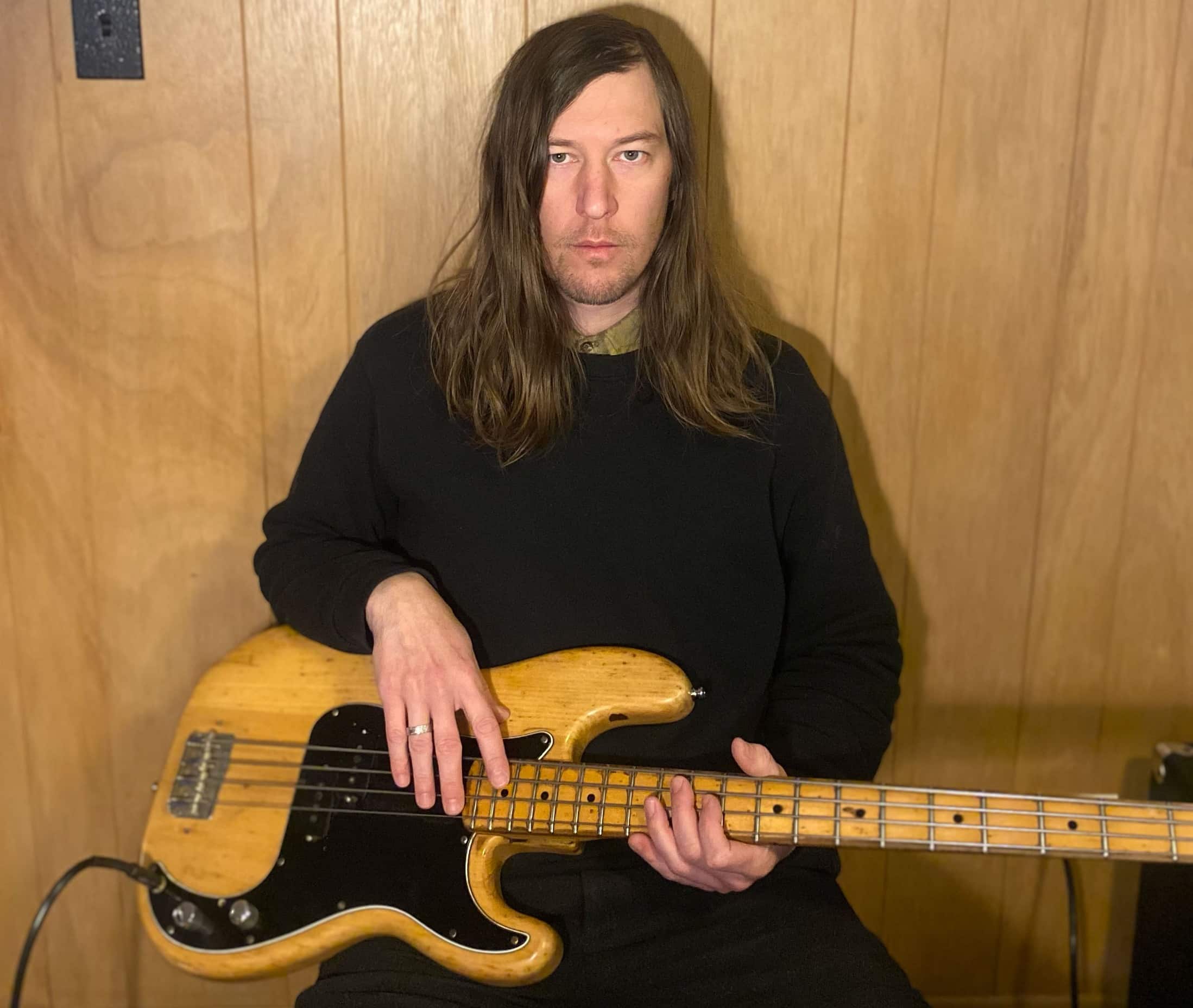 Bassist and composer Matt Ulery sits against a wood-paneled wall with his Fender electric bass in his lap. He stares forward at the camera with a neutral, stone-faced expression. Ulery wears an olive green-colored collared shirt that peeks out slightly from the top of his black sweater.