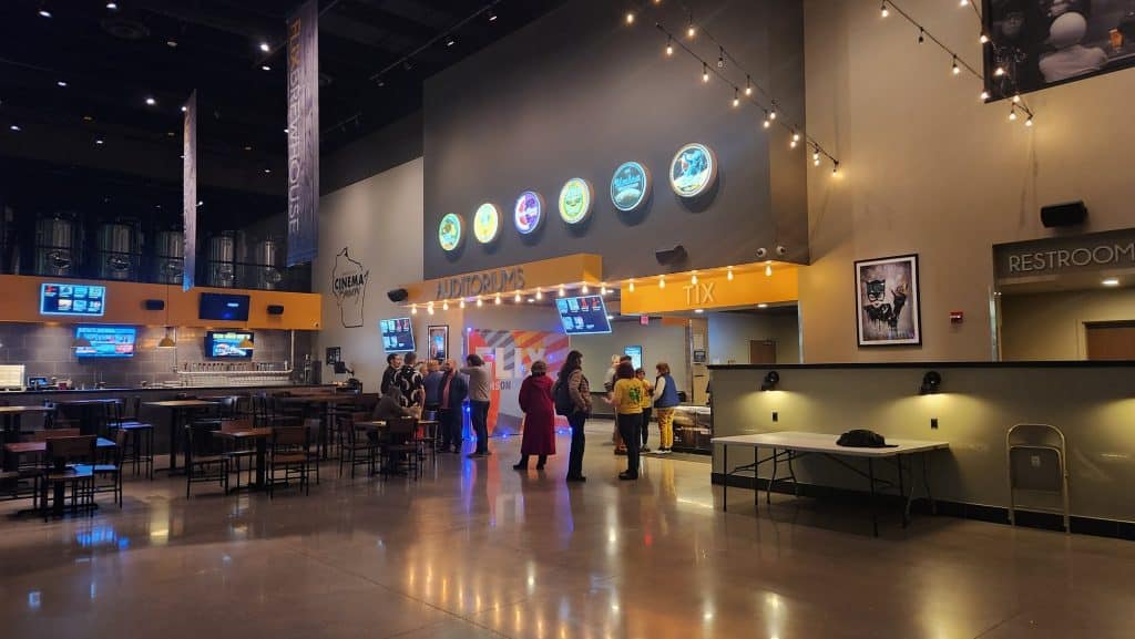 The main lobby at Flix Brewhouse at night. A photo captures the fluorescent lights of the background marquees reflecting on the marble floor in the foreground. About 10 people are gathered in small groups in the background near the "Auditoriums" sign above the entrances to the theaters.