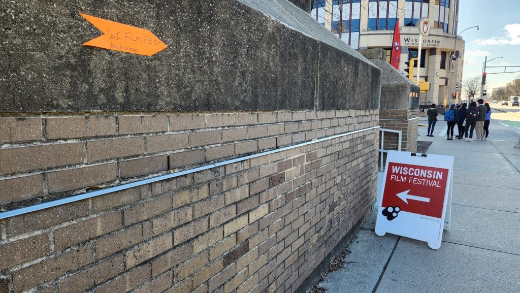 A photo on the University Avenue sidewalk near the road's intersection at North Park Street. A white sidewalk sign with the Wisconsin Film Festival logo rests against the base of Vilas Hall pointing attendees in the direction of the building's 4070 screening room. In the foreground, another small orange sticker sign on the wall itself points pedestrians towards the sidewalk sign. In the background, a red flag with the festival logo can be seen at the intersection of North Park Street, where students are standing as they await to cross the street.