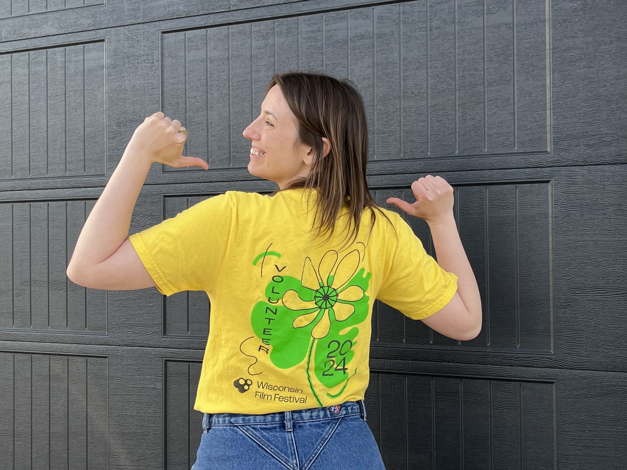The author, Sara Batkie, stands with her back of the camera in front of a slate-grey wall. She turns her head to the side, pointing with her thumbs to the green and black floral design on the back of her 2024 Wisconsin Film Festival volunteer tee shirt.