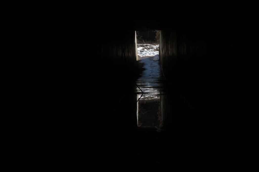 A long highway underpass is shown. The majority of the photo is pitch black though the sides of the wall can be made out. The end of the tunnel and a reflective body of water can be seen in a strip that is center-right of the image. The reflection of the outside on the water makes it appear as if there is another, upside down world. A coating of snow is visible at one of the entry-and-exit points of the underpass.