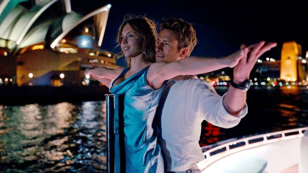 A formally dressed couple on a luxury yacht perch on the bow and reach their arms out in a reenactment of the "king of the world" scene from "Titanic" (1997). The lights of the Sydney Opera House shine behind them in the distance.