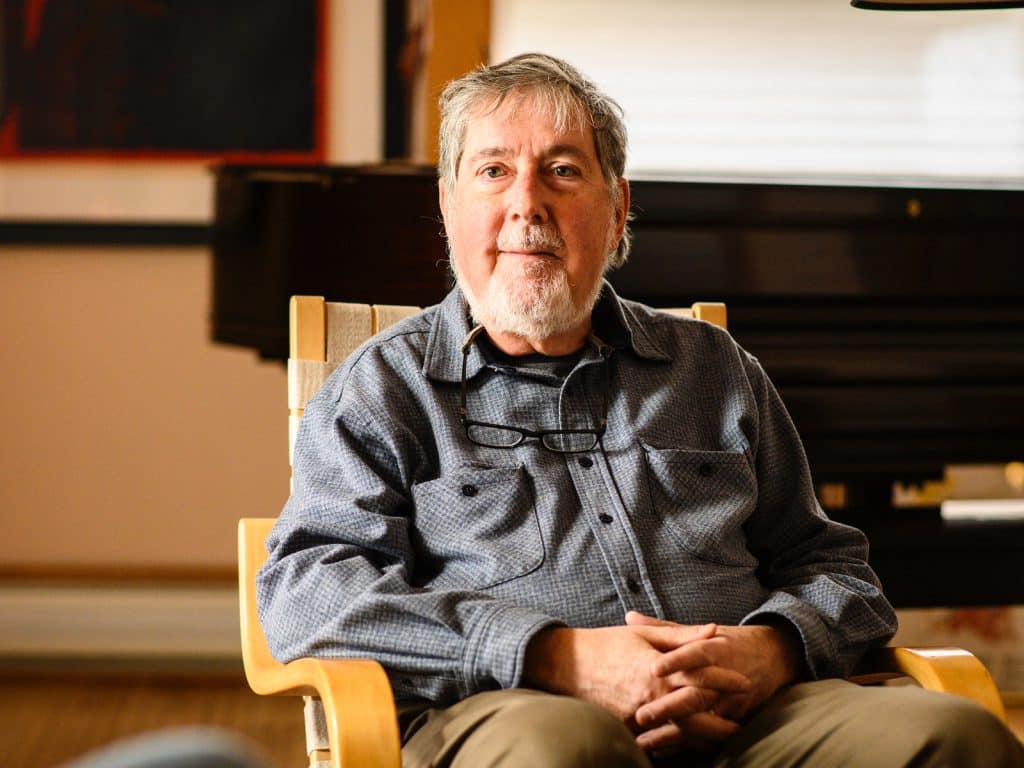 A photo shows Frank Emspak seated in a chair, facing the camera with his hands folded in his lap. In the background, out of focus, is a living room and a grand piano.