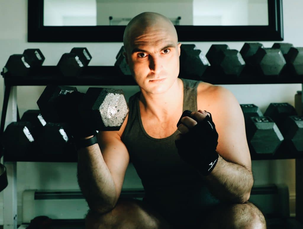 Devon Welsh poses with a small hand weight on a bench. He has the weight in mid-air, clutched in his right hand. He holds a remote light source in his left to position a bright light onto a portion of his face. He's wearing a black tank top and black fingerless gloves. Welsh is bald and muscular, staring directly at the camera. He is in the center of the image. Rows of hand weights are visible behind him.