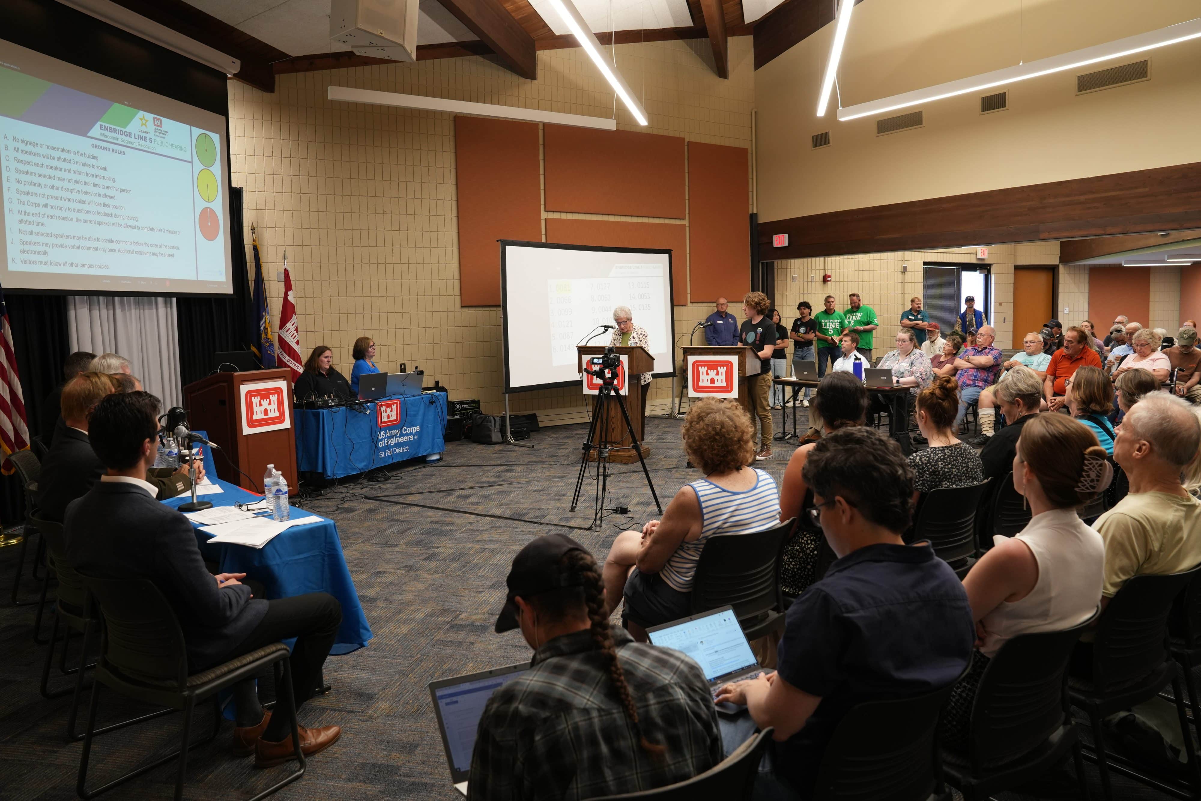 Photo of a large room with two podiums at the from with one person speaking. The audience is seated on three other sides of the room.
