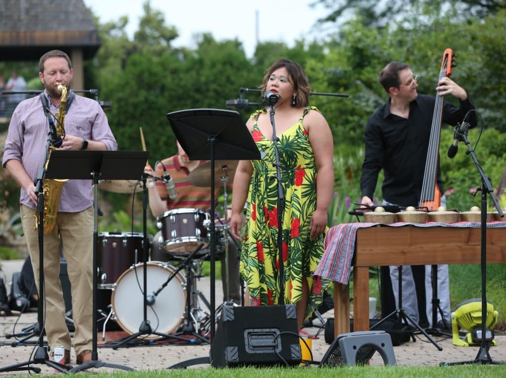 Leslie Damaso's band performs at Olbrich Botanical Gardens. From left to right: Saxophonist Luke Busch, drummer Mike Koszweski, vocalist and kulintang player Leslie Damaso, and bassist Ben Ferris. All four are in the midst of a performance in a botanical garden. In the upper left of the image, a small group is shown in extremely soft focus looking out at the performance. Everyone pictured is in casual wedding attire.