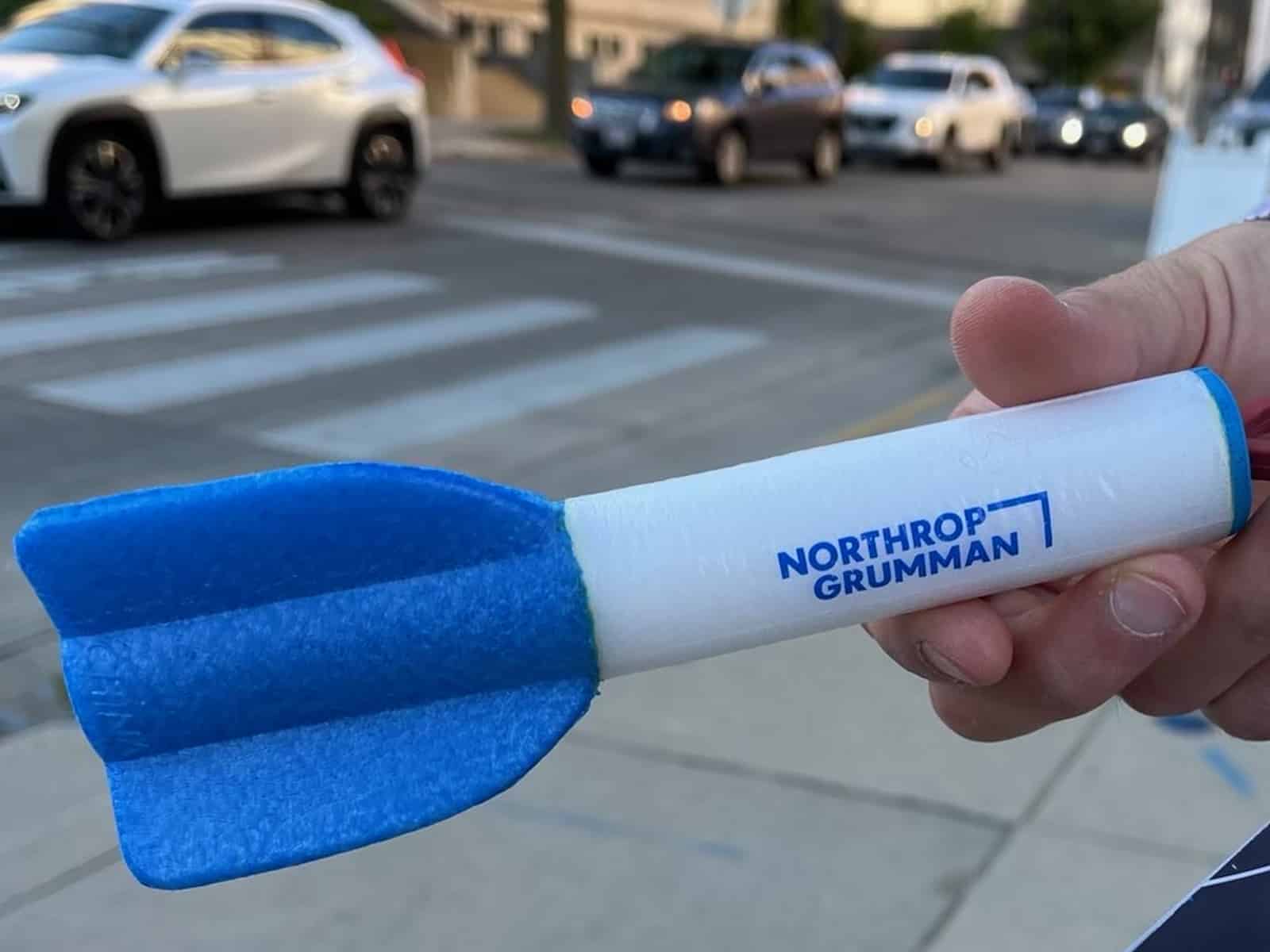 A photo shows a hand holding up a small toy missile, consisting of blue foam fins and a white plastic cylindrical body, with the Northrop Grumman logo printed on it. In the background, cars can be seen moving through a crosswalk on East Wilson Street in downtown Madison.