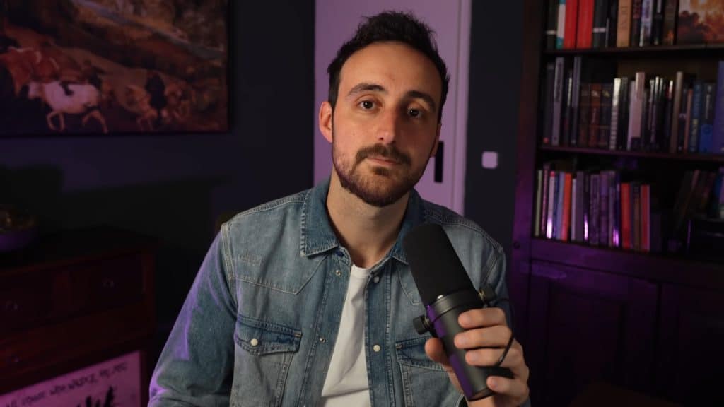 A thirtysomething man in a denim jacket sits at a close-up in an office space surrounded by a large painting behind him and a bookshelf to the right. He holds a microphone in his left hand, and stares directly at his camera, pausing in the middle of a topic.