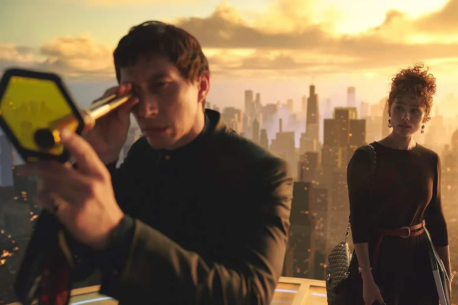Against a golden-orange CGI city backdrop, two actors stand atop a platform in dark formal attire. The man on the left peers through a spyglass at a hexagon-shaped slide in his other hand. The woman, behind him, looks over at him with intrigue.