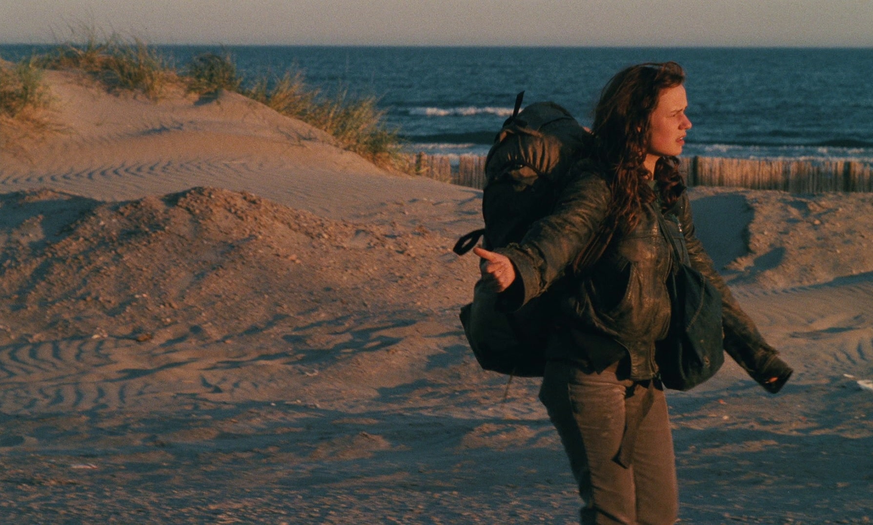 A woman in her late teens with long brown hair wears a battered leather jacket. She stands along the road near a sandy-rocky beach and coastline with her right arm and thumb pointed out towards the road in an effort to hitchhike.