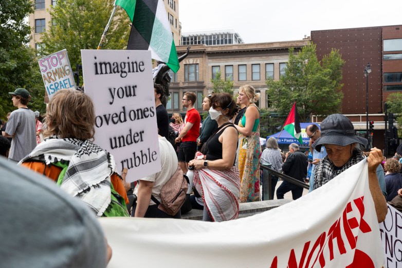 Miriam Hasan stands with her hands folded surrounded by protestors carrying banners, signs, and Palestinian flags.