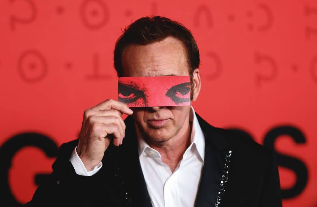 At a close-up against a blood-red background, a man in a white-collared shirt and black jacket stares forward with a neutral expression. He holds a red zoomed-in graphic of sinister-looking eyes up to his face with his right hand, covering his own eyes and most of his nose.