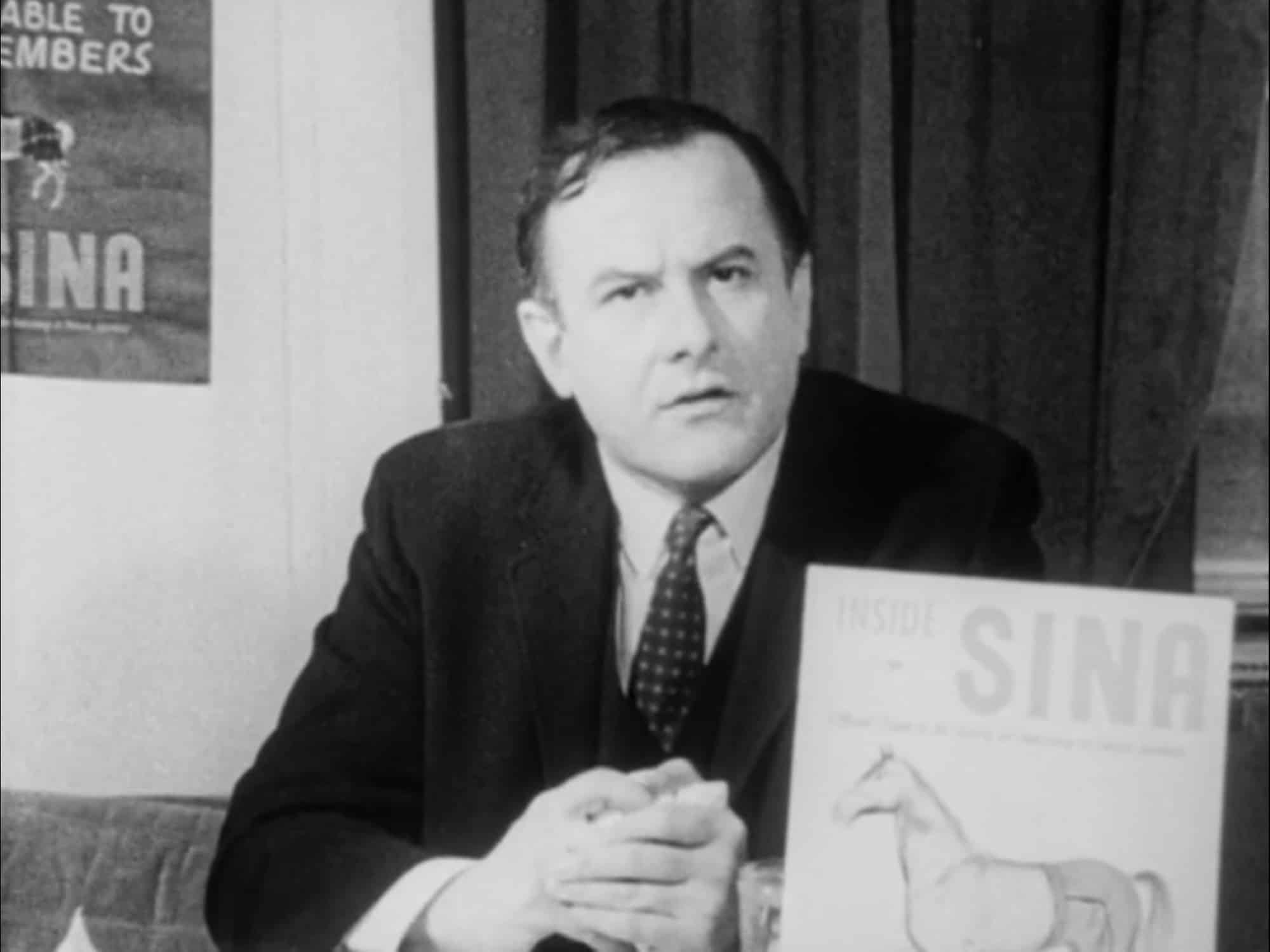 A middle-aged man in a black suit and tie stares into a camera with a serious expression. He sits at a desk with a pamphlet in front of him with a cover that reads "Inside SINA" and displays an illustration of a horse wearing pants.