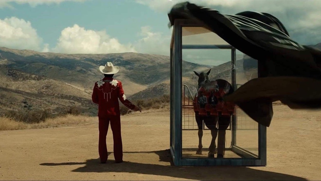 At a medium shot, a man in a red and white cowboy outfit face away from the camera, looking away out at the rolling hills and desert valley beyond. The wind blows a black sheet off a stabled horse in a cage standing to his right.