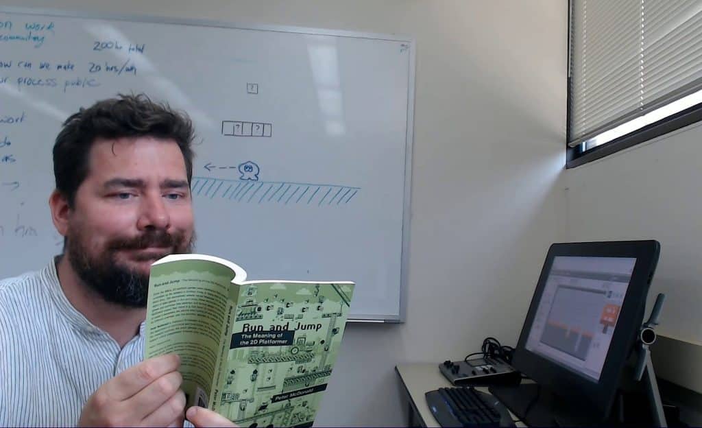 Peter McDonald, with slightly shaggy short hair and a beard, sits in an office room with a white board behind him and a computer display to his right. He playfully smirks while holding up and opening a copy of his paperback book, "Run And Jump" with a green cover illustrated by Luke Smith.