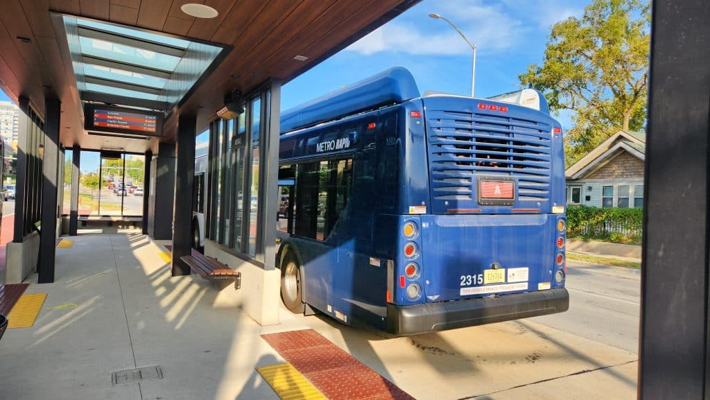 A photo shows a blue city bus leaving a bus rapid transit station. The photo is taken from inside the station. Trees, houses, and sky are visible in the background.
