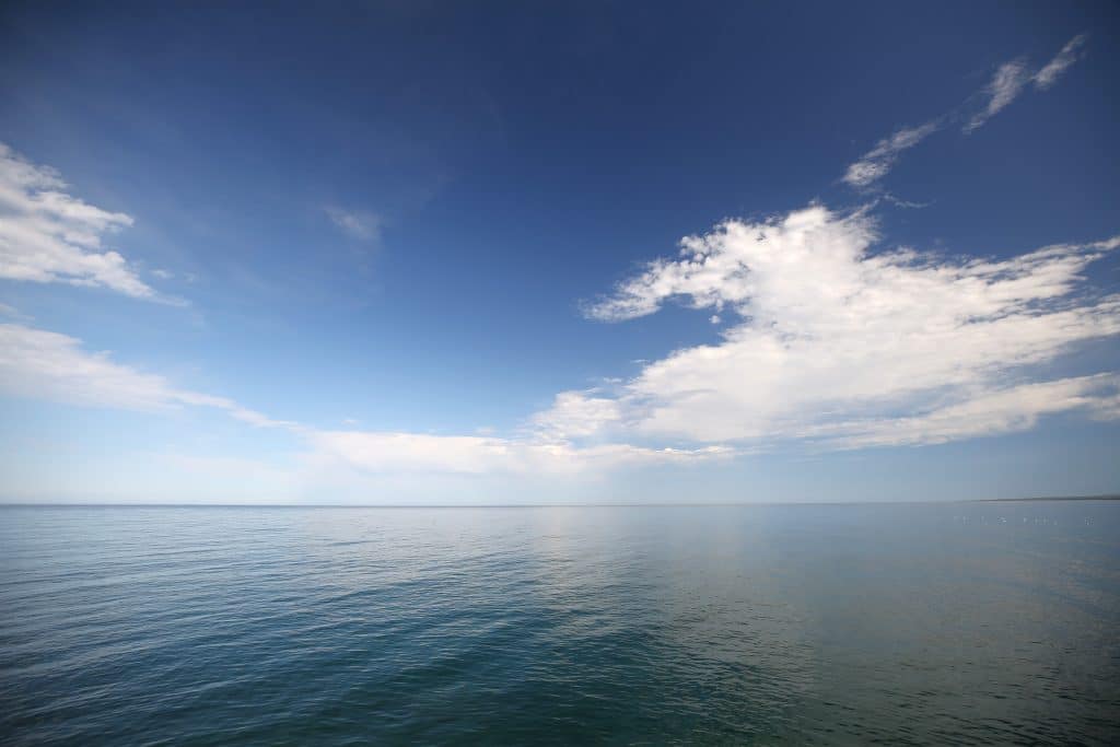 A wide-angle shot shows the sprawling expanse of Lake Superior. White clouds and a deep blue sky reflect off the lake's surface. No building are visible on the horizon, just water. On the far right of the image is a flock of seagulls floating along the water. A strip of land is also visible in the distance on the image's right side.