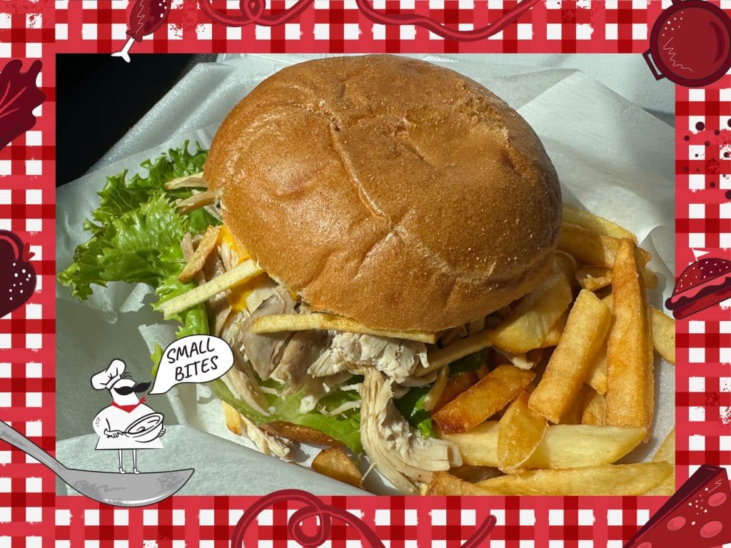 Photo of a pulled chicken sandwich, topped with yellow pepper sauce, lettuce, and fried potato sticks. An illustrated frame around the photo depicts a checked tablecloth pattern with various items of food. In the bottom left corner, a small caricature chef stands on a spoon with the speech bubble, "Small Bites."