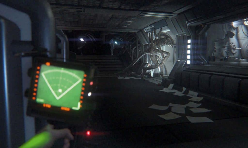 A still from a 2014 video game shows a first-person perspective of a character holding a tracking radar in front of them with their left hand. In front of them in the distance is a darkened spacecraft corridor that leads into a fluorescent-lit "Analysis Lab" hallway to the right. A black xenomorph creature is seen lurking into that hallway.