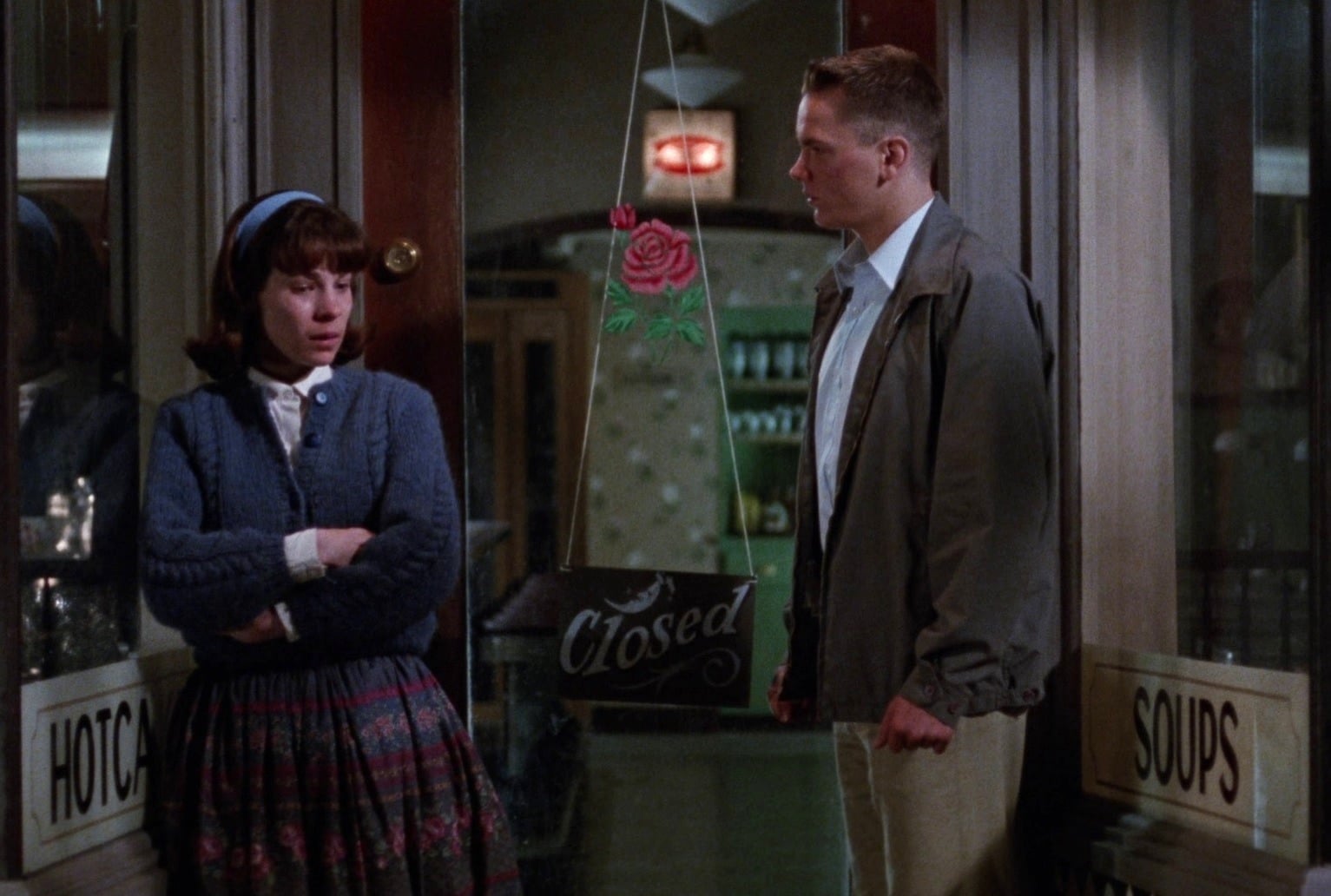 Two people in their late teens stand outside a closed shop on the streets of 1960s San Francisco at night. The young man on the right stands with his arms at his sides, looking remorseful at the young woman on the left, who is sobbing with her arms crossed. Both are semi-formally dressed.