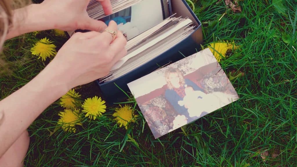A pair of hands flip through a small box of photographs resting in bright-green grass. One of the 4x6 photo prints lays face up on the grass. Its faded colors show a woman holding a child on her lap.