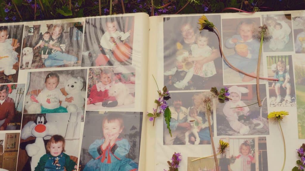 A family photo album lays open in the grass, its pages open with plucked dandelions and wild violets resting on top. The photos protected by the album's layer of plastic show the filmmaker as a baby with her mother from the mid-late 1990s.