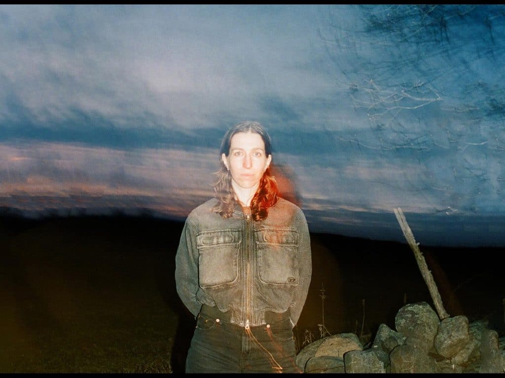 Elori Saxl stands with her hands behind her back. She is in an open field. A sunset can be seen in the distance. To the right of the image are a bare tree and a broken branch nestled into a pile of large rocks. She's wearing denim jeans and a denim jacket, and staring directly at the camera. The photograph is intentionally blurry. There are two thin black horizontal borders above and below the image.