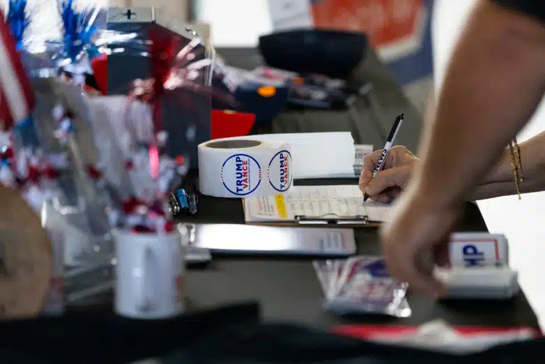 An anonymous hand writes their information on a clipboard next to Trump/Vance stickers.