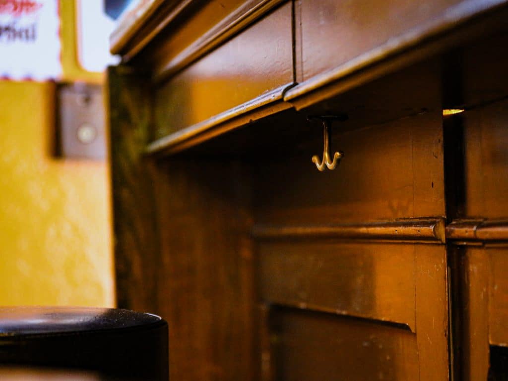 A bronze-gold bar hook is shown underneath the wooden bar at The Malt House, a bar on East Washington. The hook is in focus, in the upper right quadrant of the photograph. The bar's yellow wall and the top of a bar stool in soft focus on the left-most strip of the image.