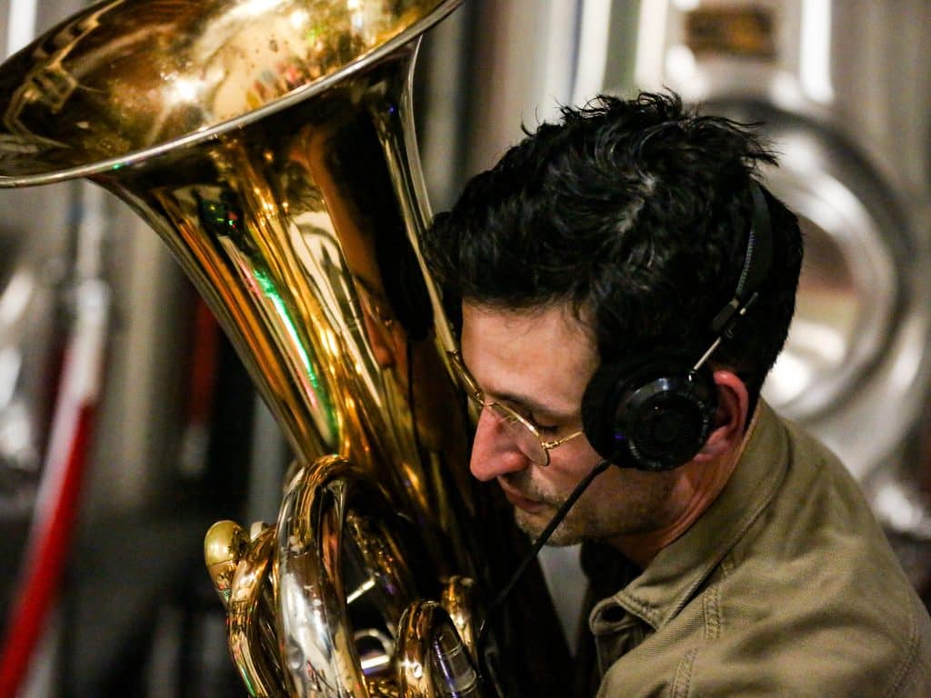 Wonderporium's Tom Curry plays tuba at Giant Jones Brewing Company for the release show of "Two Impromptus." He's wearing a muted army-green jacket, glasses, and holding his tuba close to his face. The image is a hard close-up, with the instrument cutting out of the frame on the left side of the image. Curry appears to be in a state of deep focus.