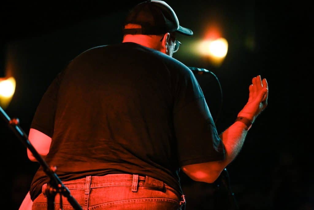 Thax Douglas reads poetry at The Majestic. He takes up the whole image. You can't see his face, only his back and the house lights ahead of him, as he holds out an arm towards the audience. He's wearing jeans, a black shirt, and a baseball cap. The photograph is taken from several feet behind him.