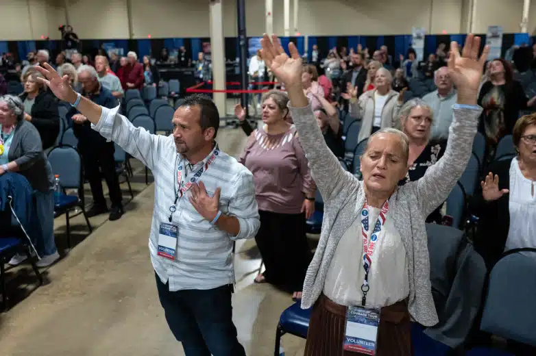 A room of people are shown engaging in a prayer service. The man and woman in the front row have their hands raised and the woman has her eyes closed.