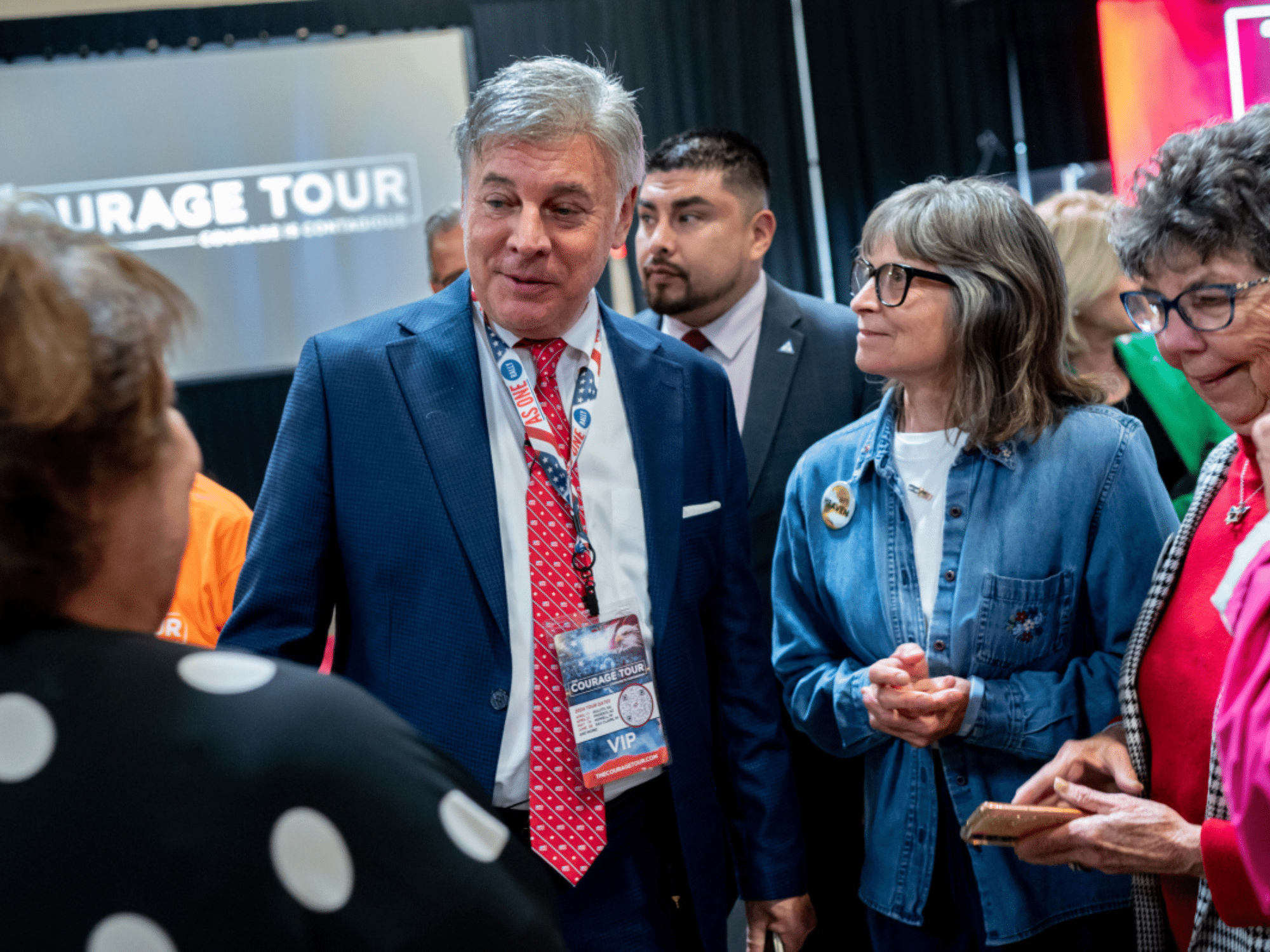 An older man with greying hair wearing a VIP badge for "The Courage Tour" greets a group of people.