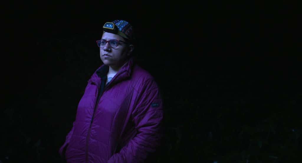 Against a black background at night, a woman in a violet-colored coat stares off-screen to the left an an unseen moth screen. She wears a headlamp that's turned off.