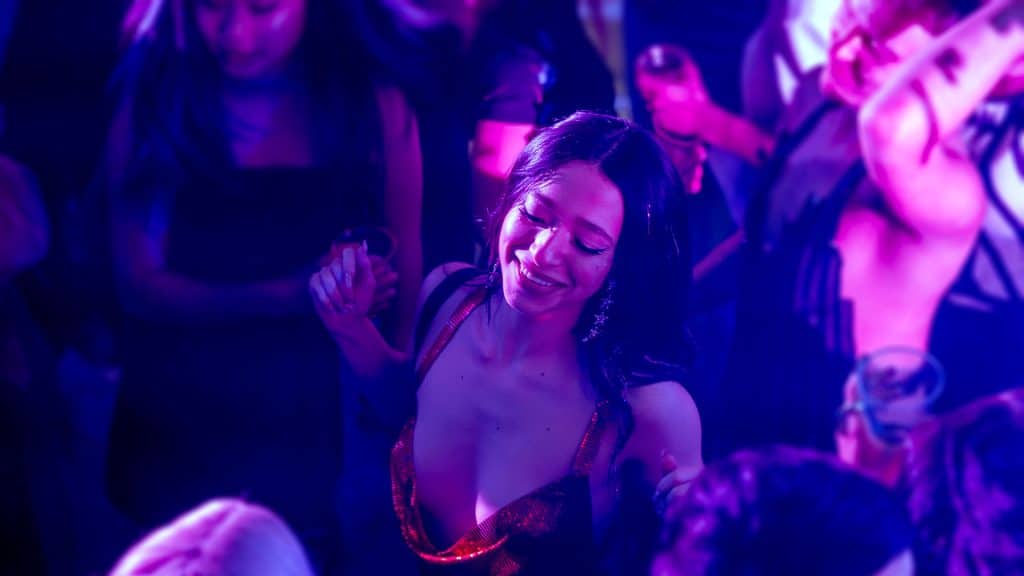 The main character of Anora is centered in the frame, dancing joyously with her eyes closed. She wears a sparkling red dress in a room with others bathed in a magenta-colored light.