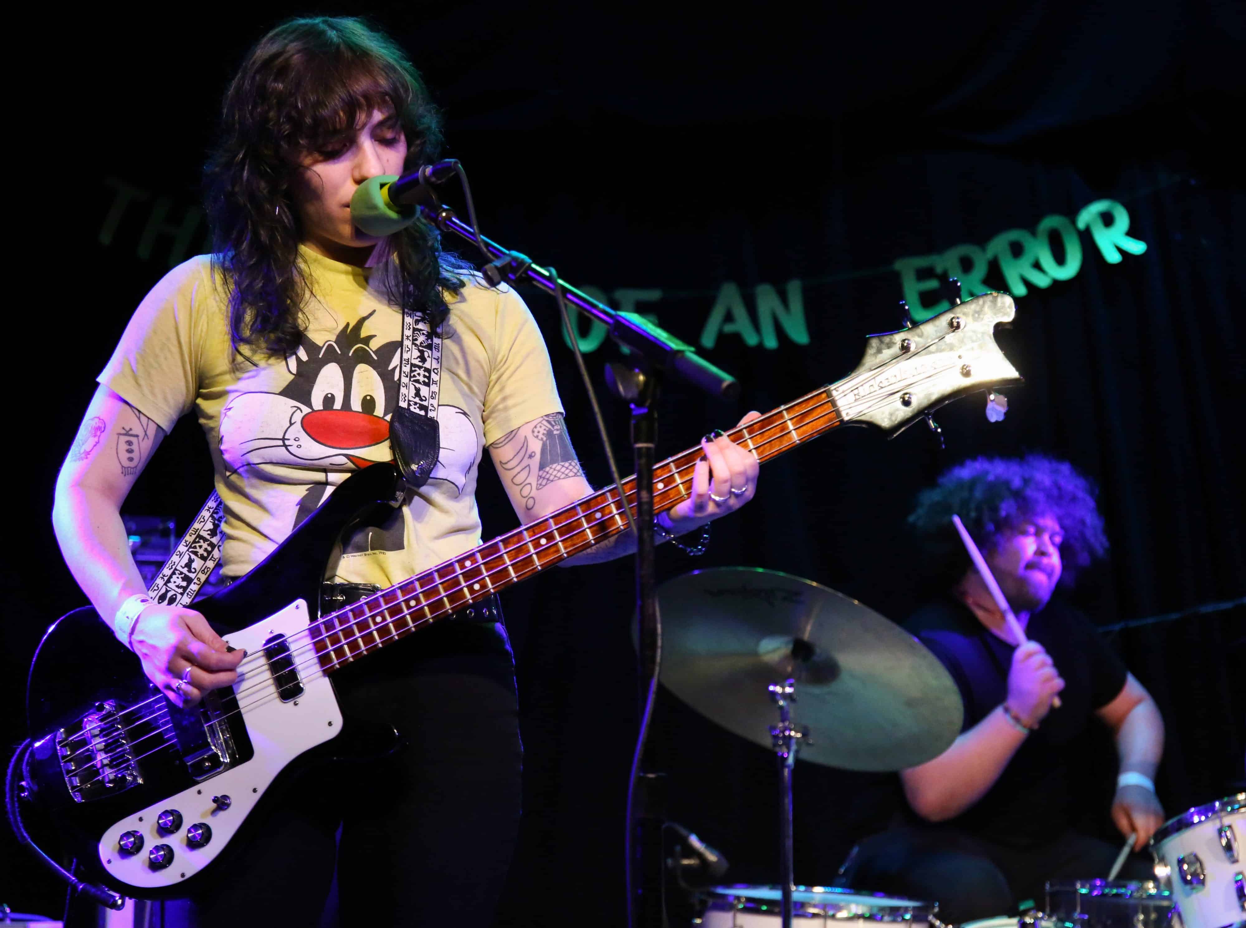 Cult Of Lip perform at High Noon Saloon. To the left of the image, Hannah Porter plays a black Rickenbacker and sings. In the lower left, Terrance Barrett plays drums. Behind them a banner that reads "The end of an error" is partially visible.