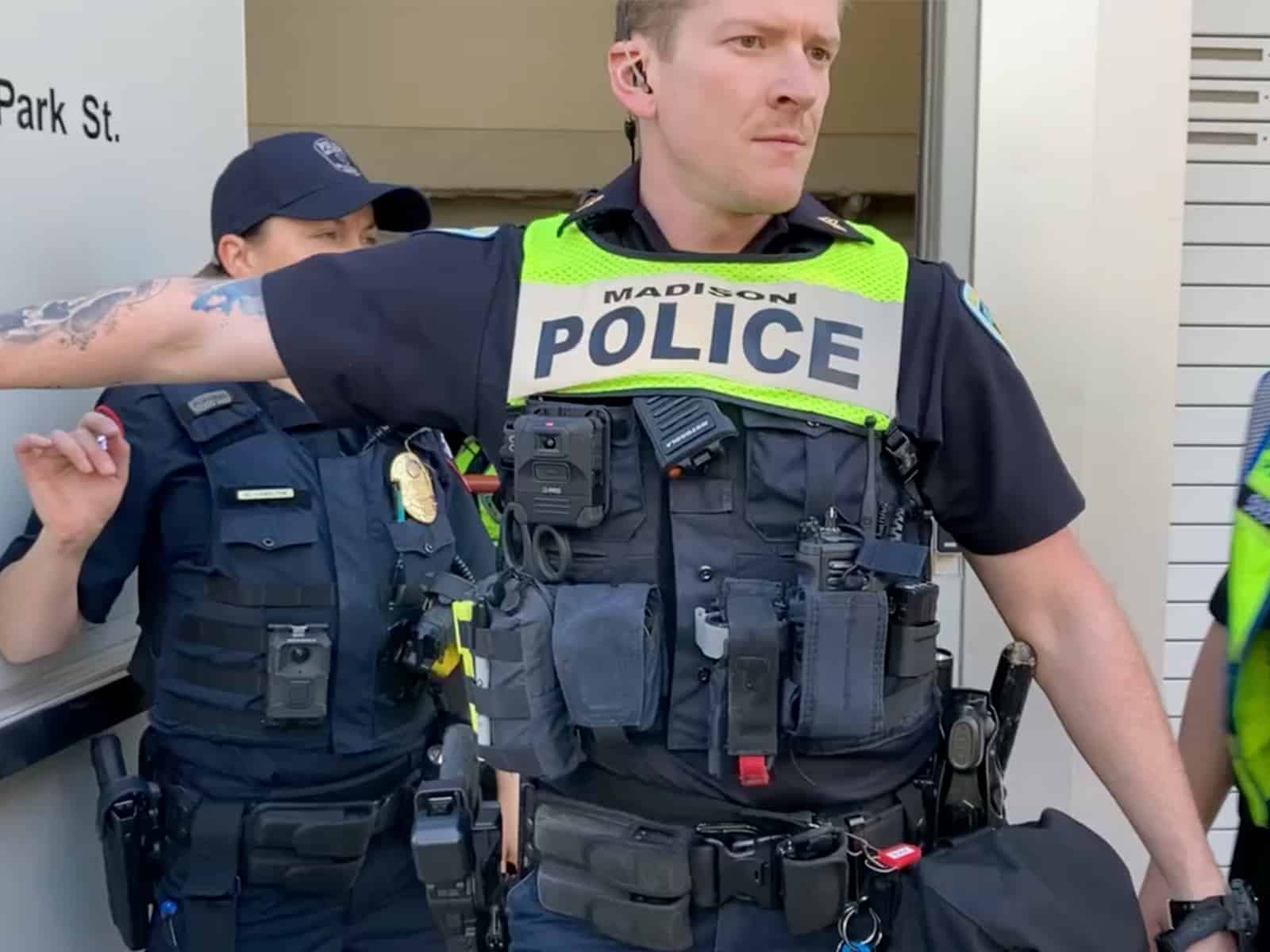 A photo shows a male police officer in patrol uniform, wearing a green mesh vest that reads "MADISON POLICE," standing outside a door and holding it open. Visible on his shirtfront are a body camera and various weapons and devices.