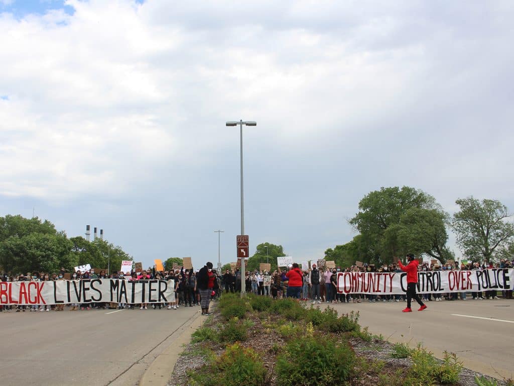 A photo shows a large group of protestors spread out across both sides of a wide road, holding two banners. One reads "Black Lives Matter" and the other reads "Community control over police."
