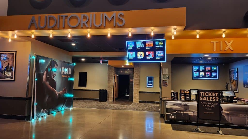 An empty movie theater lobby is illuminated by fluorescent lights above TV screens that list the eight movies currently playing at the venue. Against the left wall is a promotional banner for "Joker: Folie À Deux" that is illuminated around the edges by blue lights.